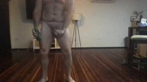 Media: Video of a naked man with a large penis, holding a green can, standing in a dimly lit room with wooden floors, a lamp, and cluttered furniture.