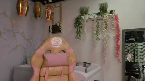 Media: Video of a pastel-colored gaming chair in a minimalist, modern bathroom with a white bathtub, pink and white floral garlands, and a small wooden shelf holding a plant.