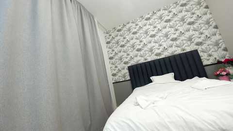 Media: Video of a modern bedroom featuring a gray upholstered headboard, white bed linens, and a wall with monochrome floral wallpaper.