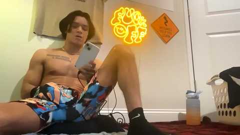 Media: Video of a shirtless man with tattoos, wearing colorful shorts and black socks, sitting on a floor, holding a phone, neon light on wall, laundry basket, and cleaning supplies in background.