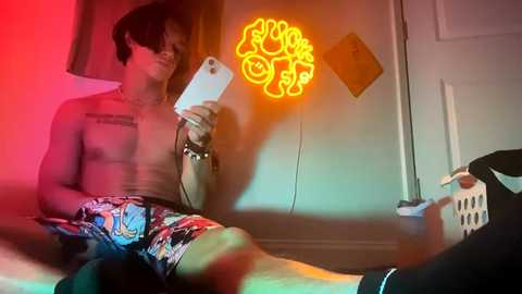 Media: Video of a shirtless, slim Asian man in colorful shorts, holding a phone, against a neon light wall, in a dimly lit room with red and yellow hues.