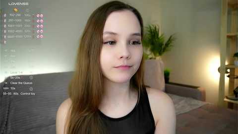 Media: A video of a young Caucasian woman with long brown hair, fair skin, and a slight smile, wearing a black sleeveless top, in a modern, softly lit living room with a gray couch, potted plants, and a lamp.