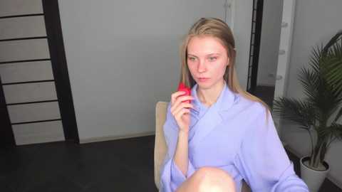 Media: Video of a blonde woman in a light blue robe, sitting on a wooden chair, holding a red lipstick. Background features a modern, minimalist room with a potted plant and a black wall.