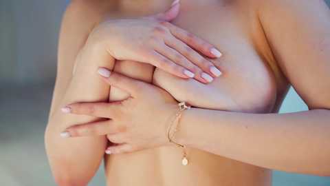 Media: Video of a fair-skinned woman, topless, covering her small to medium-sized breasts with both hands. She wears a delicate gold chain bracelet on her left wrist. Background is a blurred, neutral-toned room.