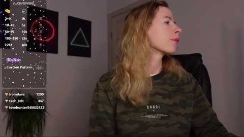 Media: A video of a young woman with light skin and shoulder-length blonde hair, wearing a camo-patterned sweatshirt, seated in a dimly lit room with a green plant and wall art.