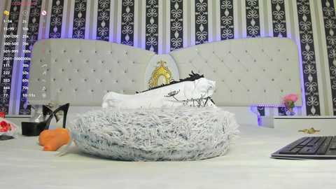 Media: A video of a luxurious, modern bedroom with a tufted white headboard, patterned curtains, and a plush grey fur rug. On the bed, a white pillow with black lace trim and a small pink rose.