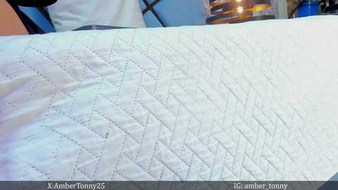 Media: Video of a neatly made, quilted white bedspread with a diamond pattern, featuring a blurry background with a blue wall and a lamp emitting warm light.