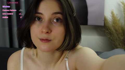 Media: A video of a young woman with short, dark hair, fair skin, and blue eyes, wearing a white spaghetti-strap top, captured indoors with a blurred background.