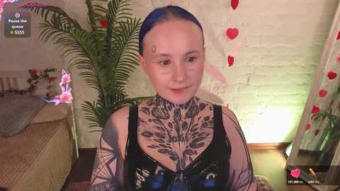 Video of a fair-skinned, blue-haired woman with intricate tattoos covering her chest and arms, wearing a black crop top, in a cozy room with a white brick wall and potted plants.