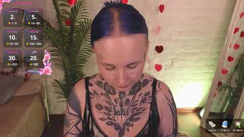 Video of a fair-skinned woman with blue hair, intricate tattoos covering her chest and arms, wearing a black sheer top, in a cozy room with a white brick wall and heart decorations.
