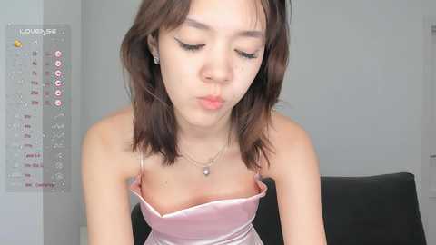 Media: Video of an Asian woman with shoulder-length brown hair, wearing a low-cut pink satin top, blowing a kiss, with a calendar and a black chair in the background.