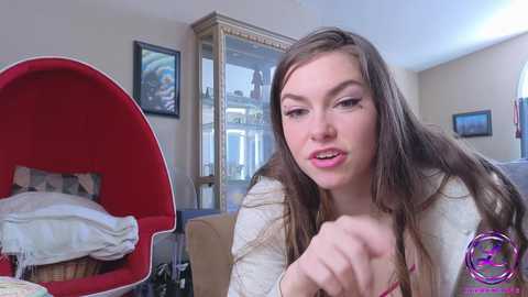 Media: Video of a Caucasian woman with long brown hair, wearing a beige cardigan, sitting in a red egg chair. Background features framed artwork, a window, and a cluttered room.