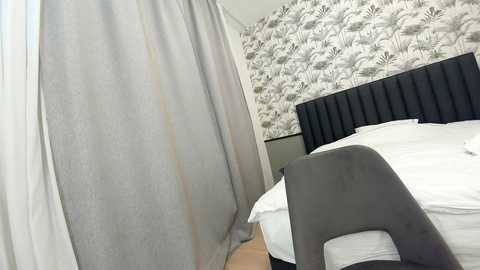 Media: Video of a modern bedroom featuring a gray upholstered headboard, white bed linens, a gray chair, and patterned wallpaper with a floral design in shades of gray and white.