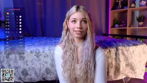 Media: Video of a young, light-skinned woman with long, wavy blonde hair, wearing a white long-sleeve top, seated on a bed with floral-patterned bedding. Background includes a bookshelf and purple lighting.