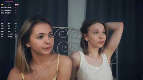 Media: A video of two young women with light skin and shoulder-length hair, wearing sleeveless tops, seated indoors with a black curtain backdrop.