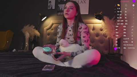 Media: Video of a young woman with long brown hair meditating on a bed with a plush, tufted headboard, wearing a floral blouse and white tights, surrounded by dim lighting, and a digital interface with text and icons.