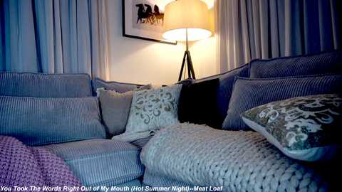 Media: Video of a cozy living room with a beige sectional sofa adorned with patterned throw pillows, a soft white knit blanket, and a tall floor lamp emitting a warm glow.
