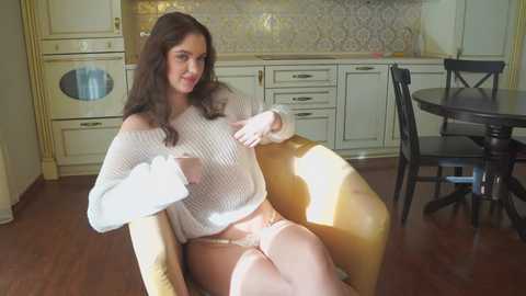 Media: A video of a woman with long brown hair, wearing a white off-shoulder sweater and yellow panties, sitting on a yellow armchair in a light-colored kitchen.
