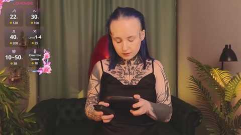 A video of a fair-skinned, tattooed woman with blue hair, wearing a black tank top and holding a smartphone, in a dimly lit room with green curtains and potted plants.