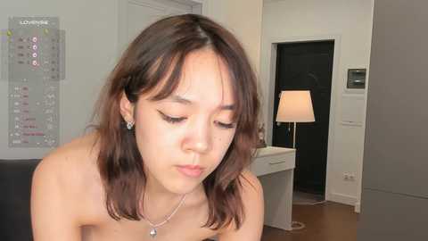 Media: Video of an Asian woman with shoulder-length brown hair, wearing a strapless top, looking down, in a modern, minimalist room with a white desk, lamp, and calendar.