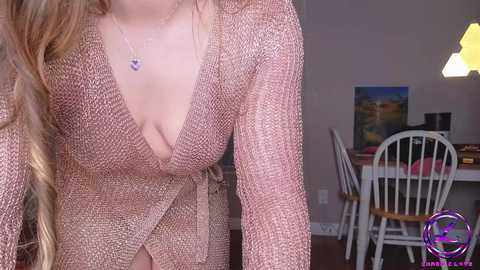 Media: Video of a woman in a pink, textured cardigan, revealing a deep V-neckline, with long blonde hair, wearing a delicate necklace. Background shows a dining room with white chairs, a painting, and a modern pendant light.