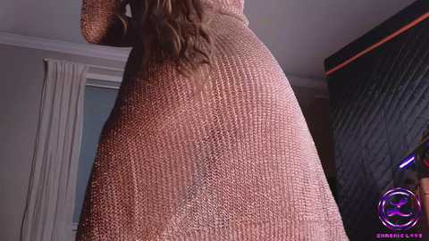 Media: A video showing a woman from behind, wearing a textured, brown knit dress that clings to her curvy, shapely hips and thighs. Her long, wavy hair cascades down her back. The background features a modern, dimly lit room with a window and dark furniture.