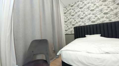 Media: Video of a minimalist bedroom with a black upholstered headboard, white bed linens, and a gray chair near a partially open curtain with a floral patterned wallpaper.