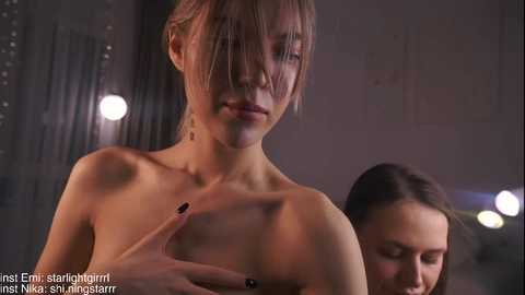 Media: Video of a topless, slender woman with light skin, shoulder-length blonde hair, and small breasts, standing in a dimly lit, industrial setting.