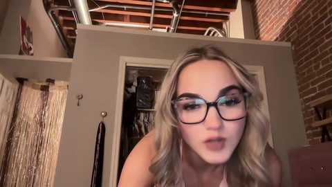 Media: A video of a young woman with long, wavy blonde hair, wearing black-rimmed glasses, leaning over a railing, with a partially open closet door in the background, showcasing a cluttered interior with exposed brick walls and wooden beams.