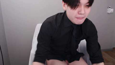Media: Video of a young Asian man with fair skin and black hair, wearing a black shirt and no pants, sitting on a white chair in a plain room with gray walls.