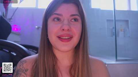Media: Video of a smiling, topless woman with long brown hair and a tattoo on her left shoulder, standing in a bathroom with a glass shower.