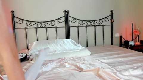 Media: Video of a neatly made bed with white sheets and pillows, black wrought iron headboard, and a partially visible person in the foreground, against a pale green wall.
