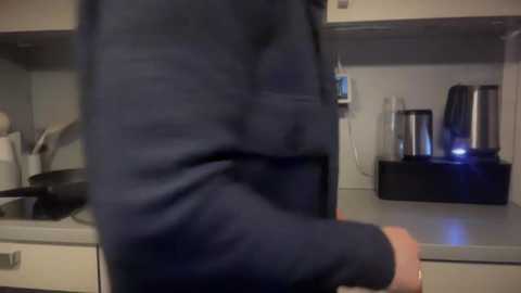 Media: A video of a person in a navy blue sweater standing in a modern kitchen. The background features white cabinets, a stainless steel kettle, and a toaster. The image is slightly blurry.