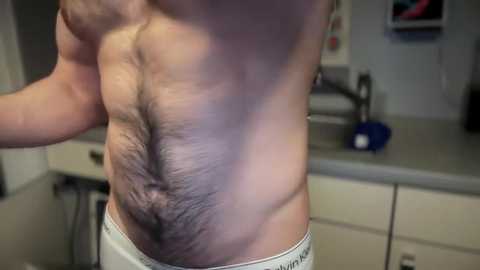 Media: Video of a shirtless man with a hairy chest and midsection, wearing white Calvin Klein boxer briefs, standing in a kitchen with beige cabinets, a sink, and a blue towel.