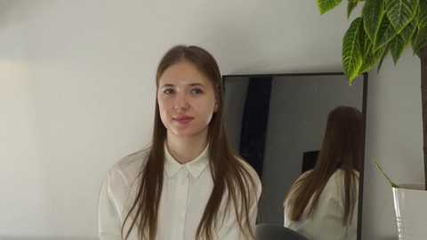 Video of a young woman with long brown hair, fair skin, and a neutral expression, wearing a white blouse, standing in a modern room with a mirror and green plant in the background.
