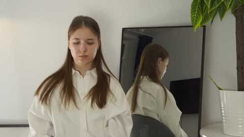 Video of a young woman with straight, long brown hair, wearing a white blouse, standing in front of a mirror reflecting her back, in a minimalist, modern room with a white wall and green plant.