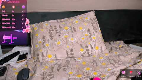 Media: Video of a bedroom with a beige pillow and bedspread adorned with yellow daisy flowers, featuring a black headboard. A television displaying a pink fox and various icons is on the left, and a smartphone rests on the bed.