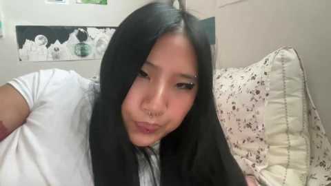 Media: Video of an Asian woman with long black hair and fair skin, wearing a white shirt and black jacket, pouting, in a bedroom with white walls and floral-patterned pillow.