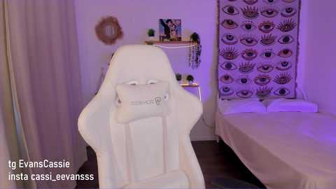 Media: Video of a serene, minimalist spa room featuring a white, ergonomic massage chair with a cushioned headrest. A bed with white linens is adorned with a decorative tapestry in the background.