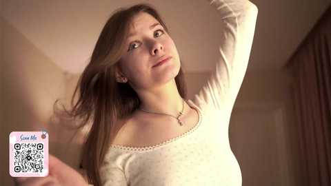 Media: A video of a young Caucasian woman with long, straight brown hair, wearing a white off-shoulder sweater, standing indoors with her head tilted back and hair held up, in a soft, warm light.