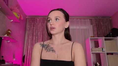 Media: Video of a young woman with light skin, brown hair, and a floral shoulder tattoo, wearing a black spaghetti strap top, standing in a pink-lit bedroom with shelves and curtains in the background.