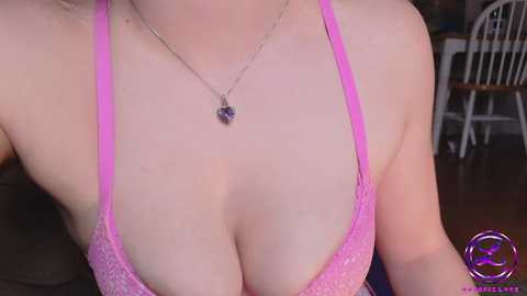 Media: A close-up video of a light-skinned woman with fair, smooth skin wearing a pink lace bra, showcasing cleavage, with a silver necklace featuring a small pendant. Background includes a wooden floor and a white chair.