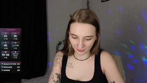 Media: Video of a young woman with light skin and straight brown hair, wearing a black tank top, standing in a dimly lit room with a digital clock displaying 10:59 PM, visible tattoos on her arm, and a black choker.