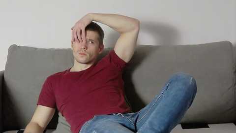 Media: Video of a muscular, fair-skinned, short-haired man with a serious expression, wearing a red T-shirt and blue jeans, sitting on a gray couch against a plain white wall.
