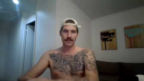 Media: Video of a shirtless, muscular white man with a mustache, wearing a backward baseball cap, standing in a minimalist room with two abstract paintings on the wall.