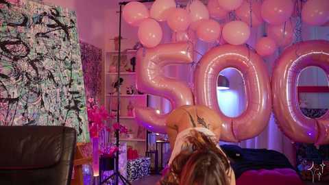 Media: A vibrant, modern room with a dark leather couch, a large abstract painting, and pink \"50\" balloons, featuring a woman with tattoos leaning forward, a pink floral table, and a zebra-striped blanket.