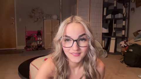 Media: Video of a young woman with long, wavy blonde hair and glasses, smiling in a cluttered, dimly lit bedroom.