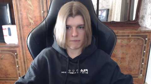 Media: Video of a young woman with shoulder-length blonde hair, wearing a black hoodie with \"A.I.R\" branding, sitting in a black gaming chair, against a backdrop of wooden furniture and a lace curtain.