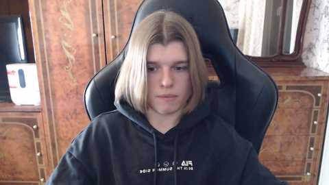 Media: Video of a young person with shoulder-length blonde hair, wearing a black hooded sweatshirt, sitting in a black gaming chair. The background features ornate wooden cabinets with intricate carvings and a television screen.