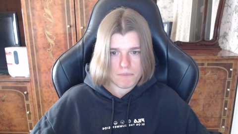 Media: A video of a young, pale-skinned man with shoulder-length, straight blonde hair, wearing a black hoodie, seated in a black gaming chair. The background features a wooden cabinet with ornate designs and a mirror.